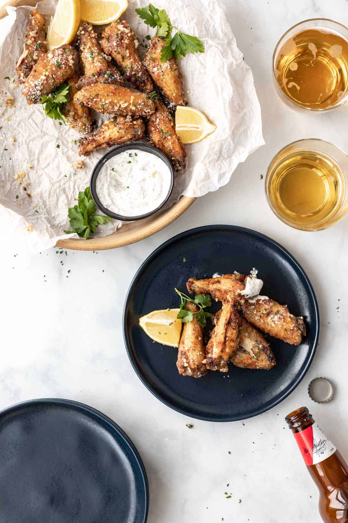 Chicken wings on a blue plate surrounded by a serving dish full of wings, 2 cups of beer, an empty beer bottle, and a bottle cap.