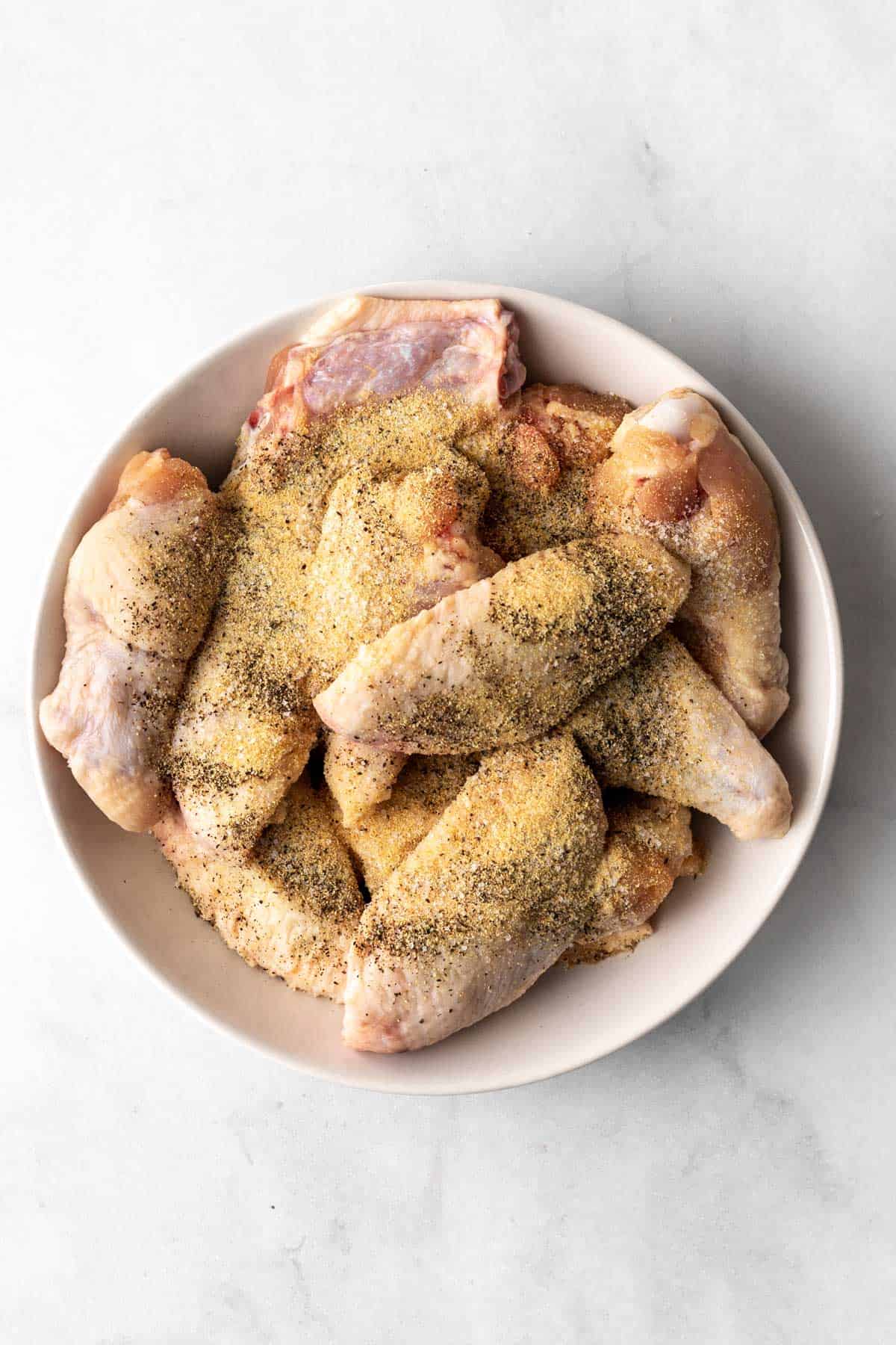 Raw chicken wings in a white bowl with salt, pepper, and garlic powder on top.