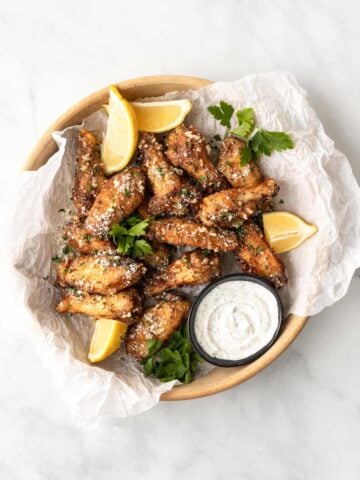 Air fryer garlic parmesan wings in a serving dish, garnished with lemons and parsley, and served with a side of ranch.