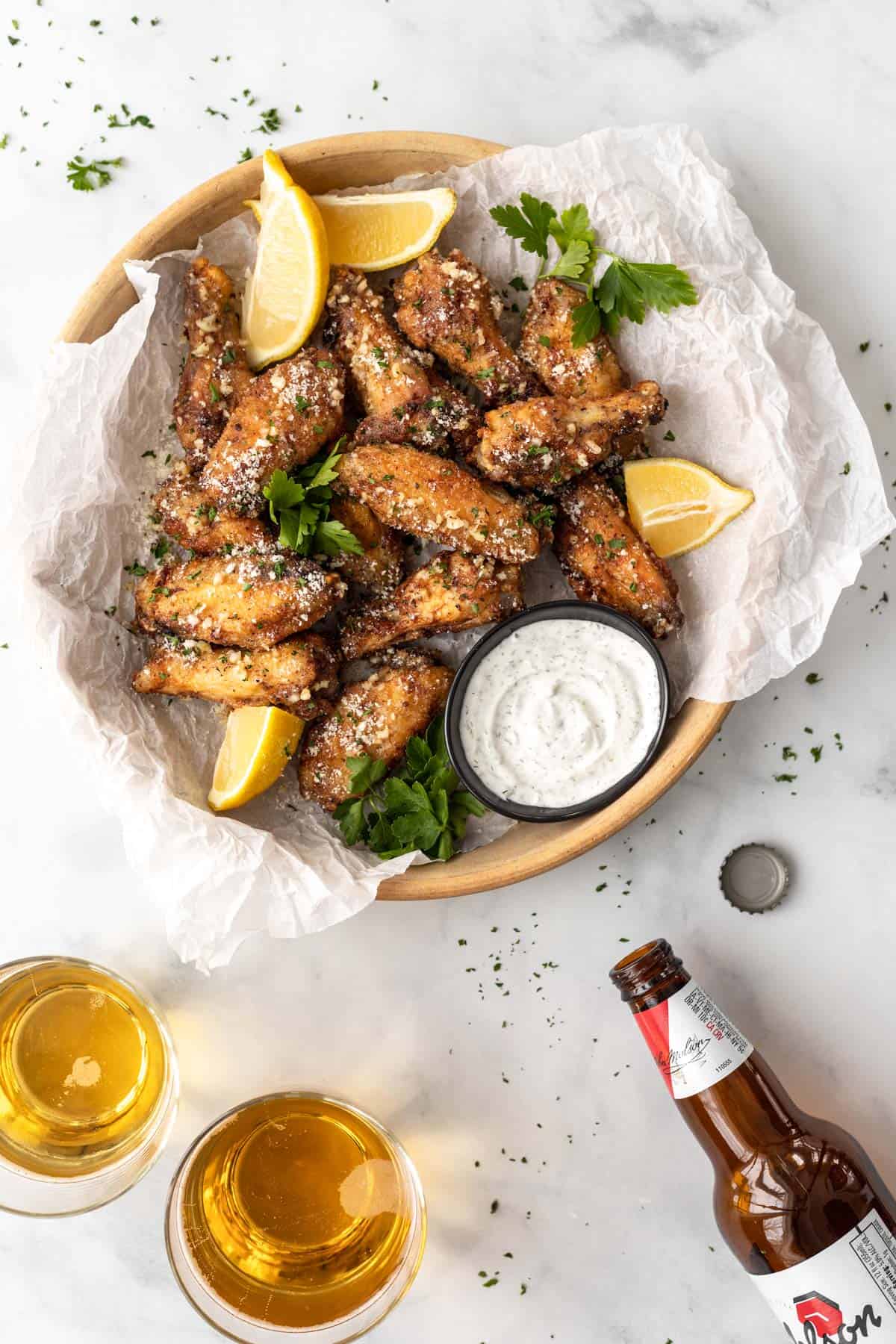 Air fryer parmesan garlic wings piled into a serving dish with a side of ranch, 2 glasses of beer, a beer bottle, and a bottle cap.