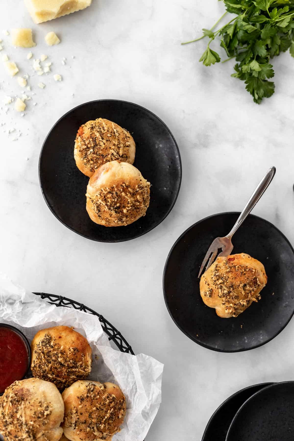 Air fryer pizza rolls on black plates with a basket of rolls, a bunch of parsley, and crumbs of cheese around it.