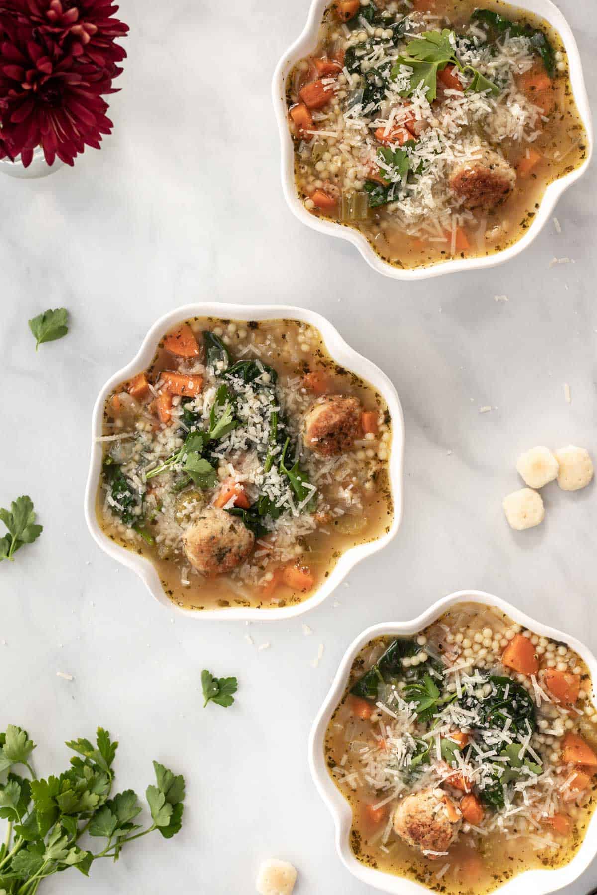 3 bowls of soup on a white backdrop with maroon flowers, parsley, and oyster crackers scattered around them.