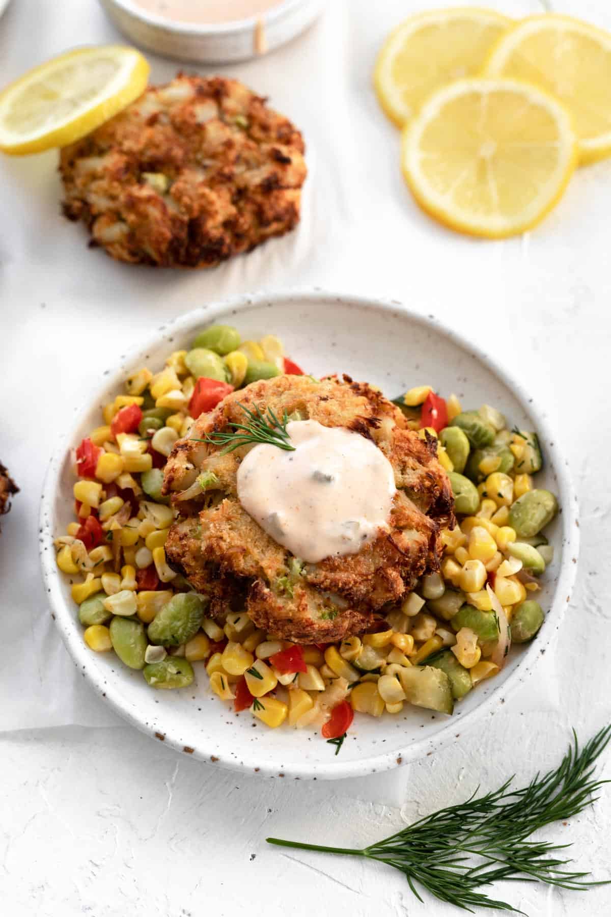 A crab cake on top of corn succotash on a white plate with a dollop of homemade remoulade sauce on top.