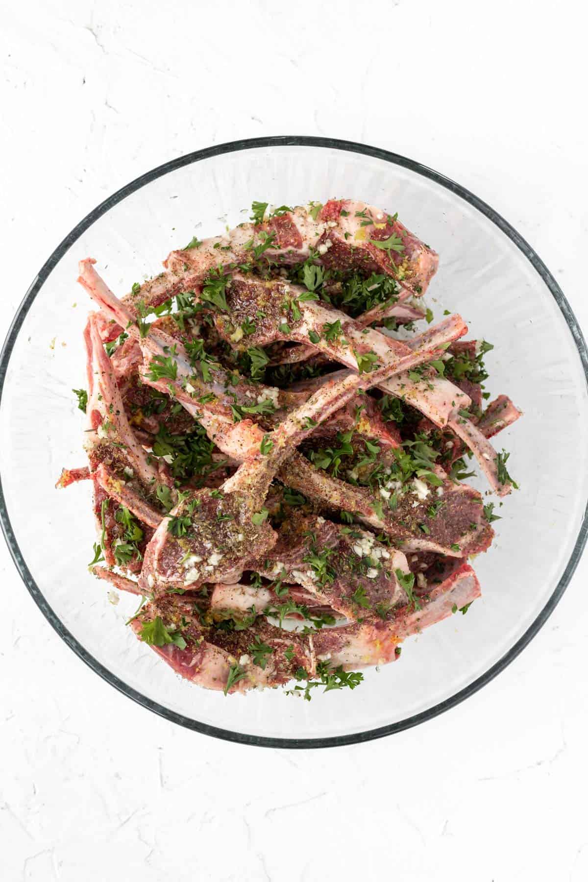 Raw lamb chops in a large glass bowl mixed with marinade.