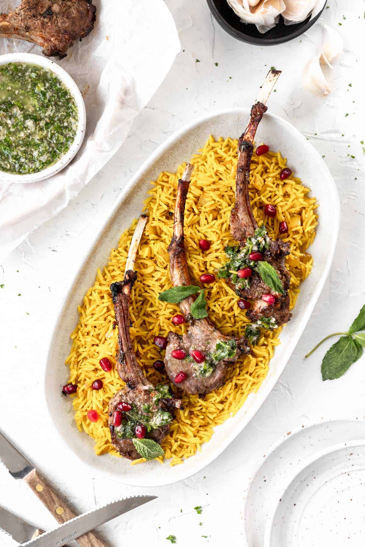 3 lamb chops on an oval platter with yellow rice, mint chimichurri, and pomegranate seeds.