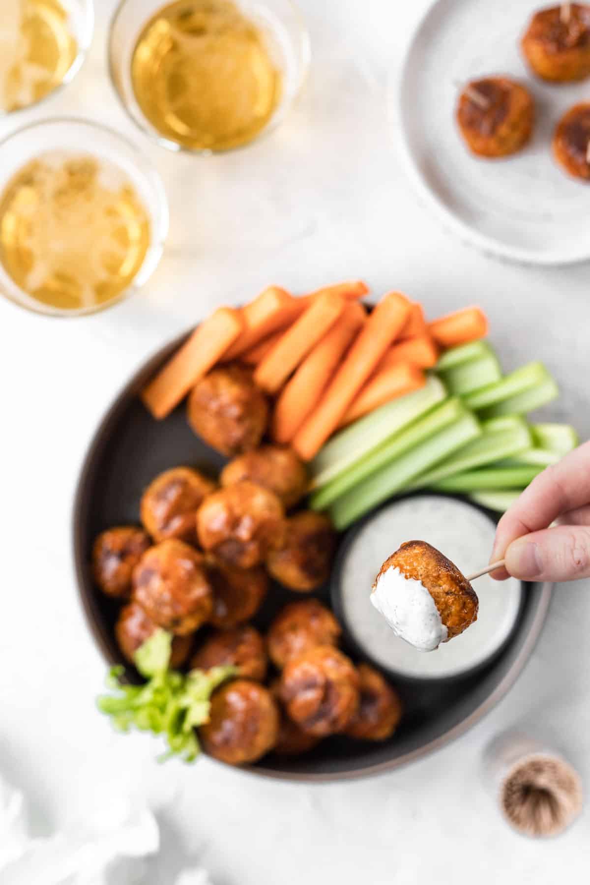 A hand dipping a buffalo turkey meatball in feta ranch with glasses of beer, celery and carrot sticks, and a jar of toothpicks off to the side.