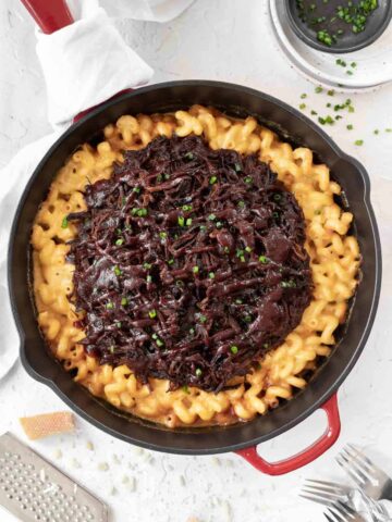 Creamy cheddar baked brisket mac and cheese in a large cast iron skillet with chopped chives and grated cheese around it.