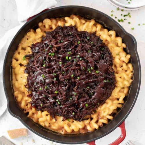 Creamy cheddar baked brisket mac and cheese in a large cast iron skillet with chopped chives and grated cheese around it.