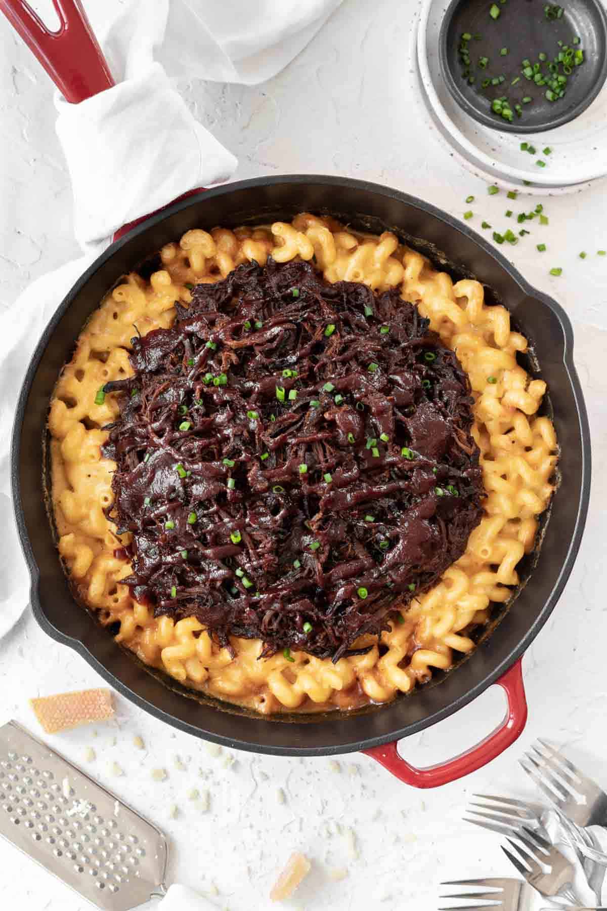 Creamy cheddar baked brisket mac and cheese in a large cast iron skillet with chopped chives and grated cheese around it.