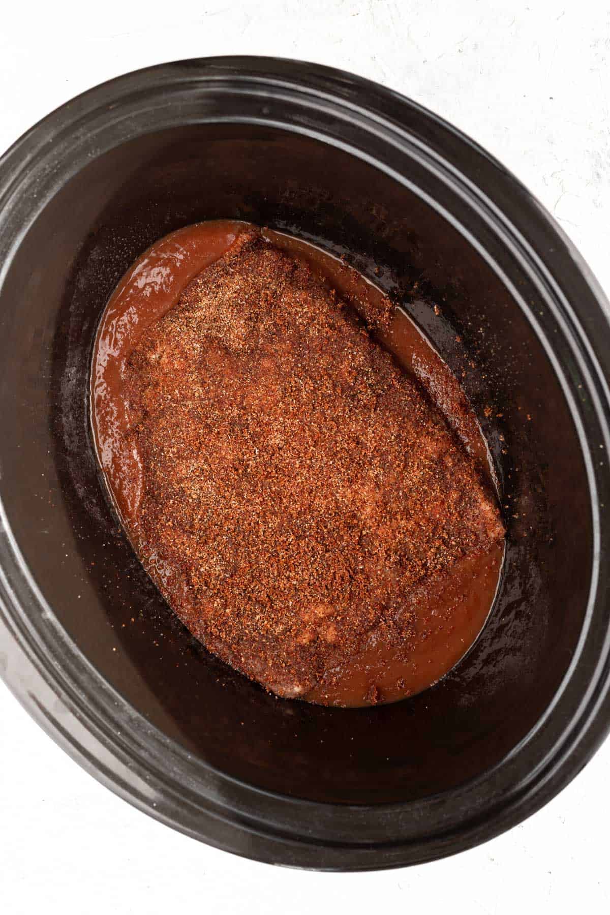 Dry rubbed brisket on top of sauce in the base of a slow cooker.