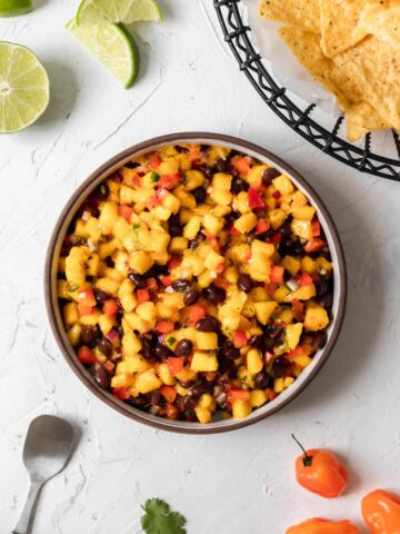 Spicy mango habanero salsa in the center of sliced limes, habaneros, a basket of tortilla chips, and a serving spoon.