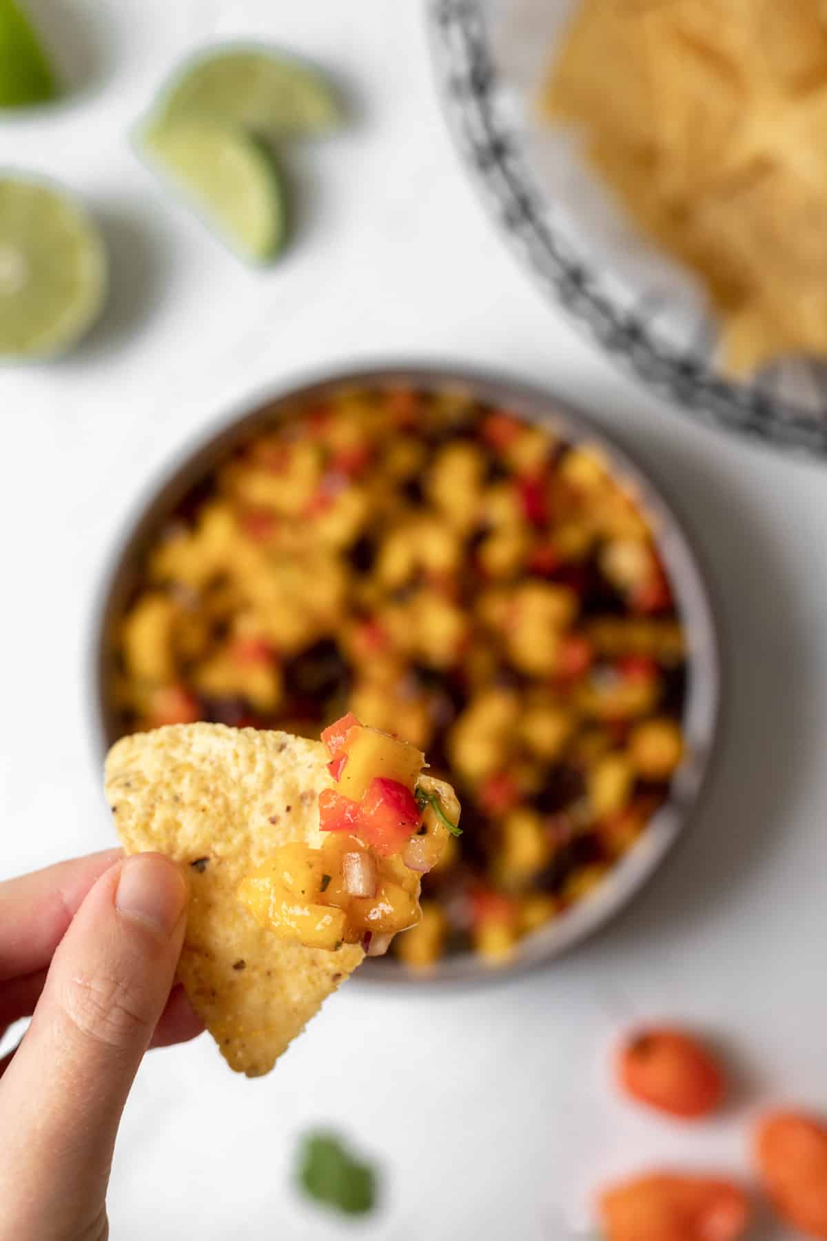 Spicy mango salsa being scooped up by a tortilla chip above and bowl of salsa and a basket of more tortilla chips.