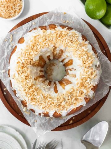 Coconut lime drizzle cake on a wooden cake stand with fresh limes around it.