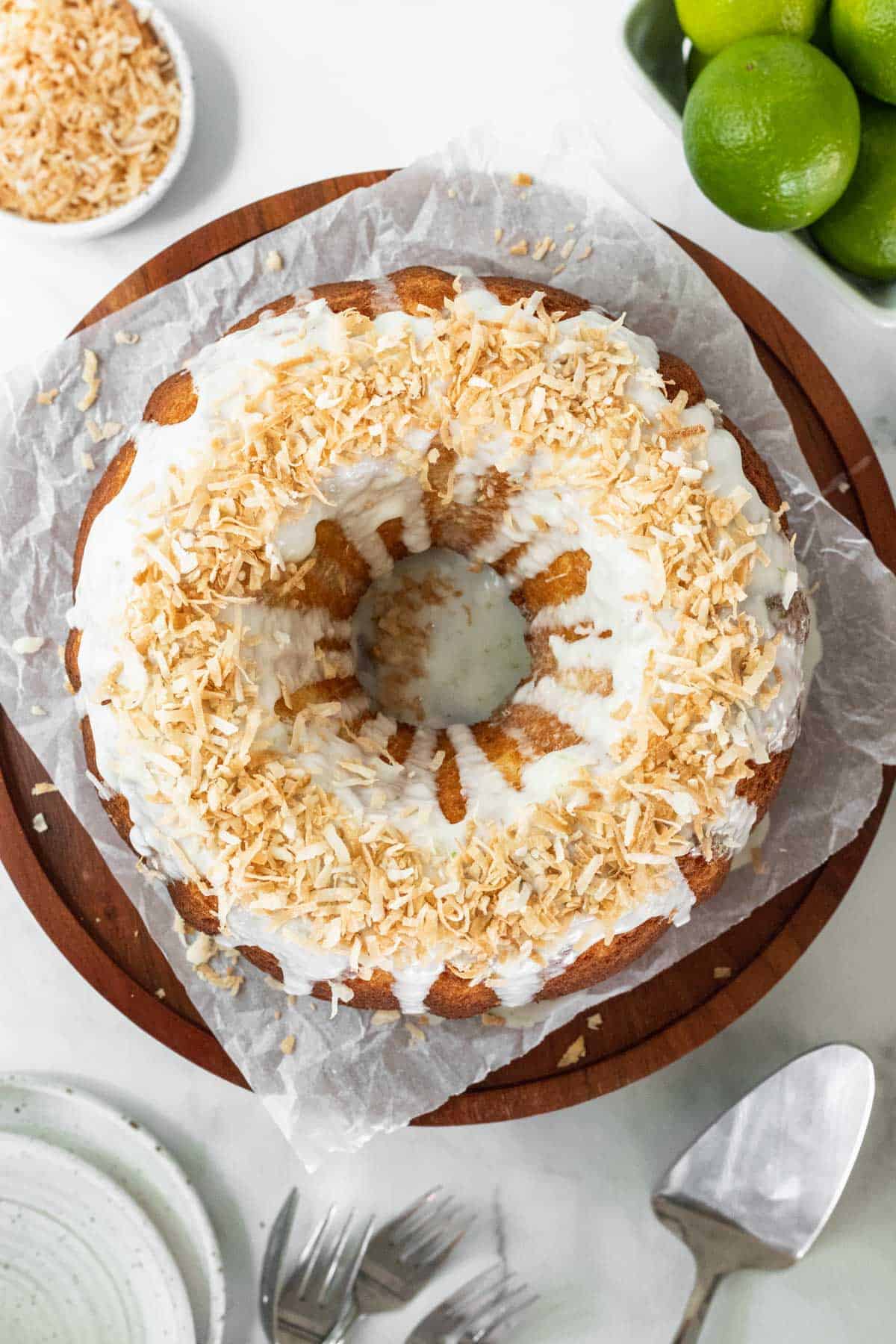 Coconut lime drizzle cake garnished with toasted coconut on a wooden cake stand with fresh limes around it.