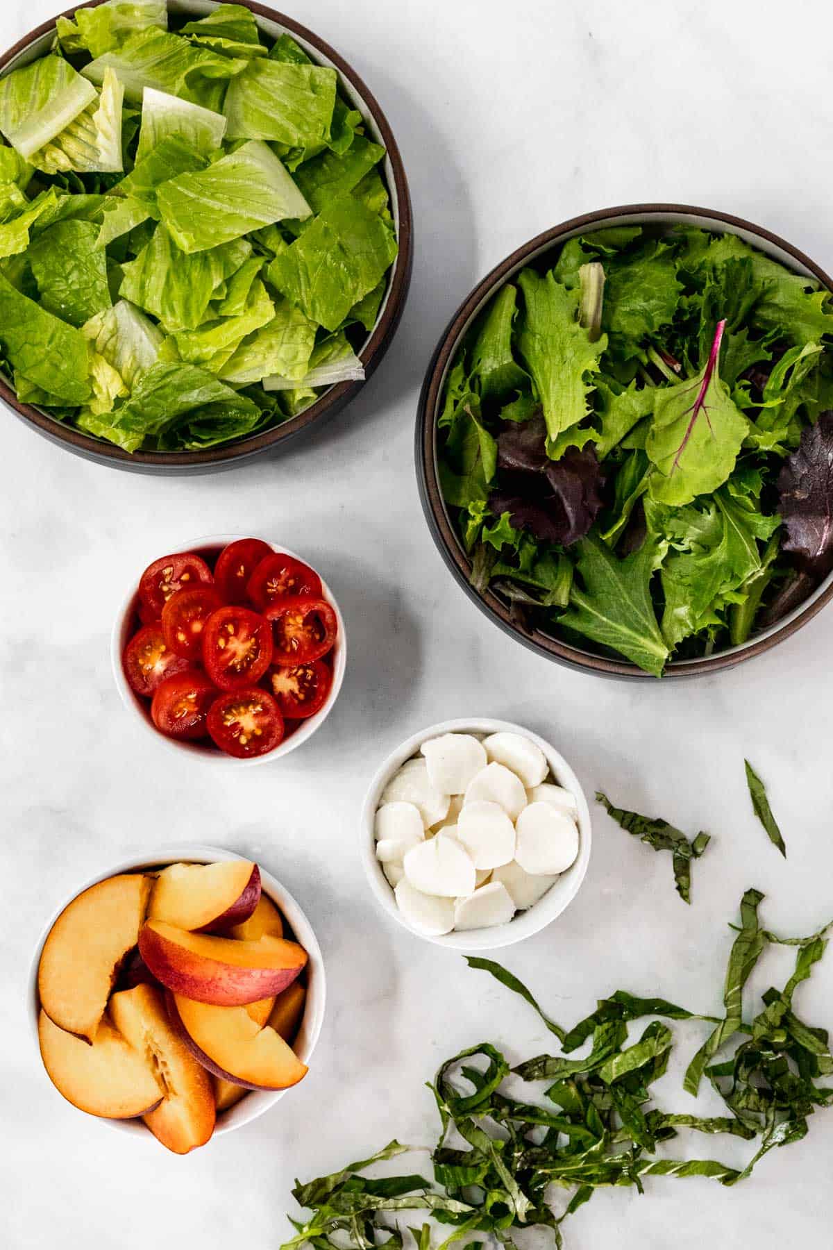 Chopped lettuce and basil, sliced tomatoes and mozzarella, and cut peaches in bowls ready to be assembled.