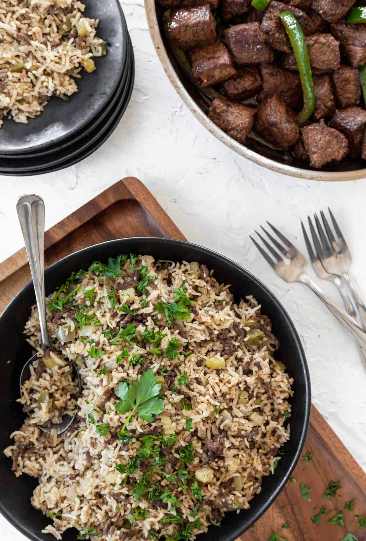 Instant pot dirty rice in a bowl with a serving spoon, a black plate with some rice on it, and a skillet of cajun steak bites off to the side.