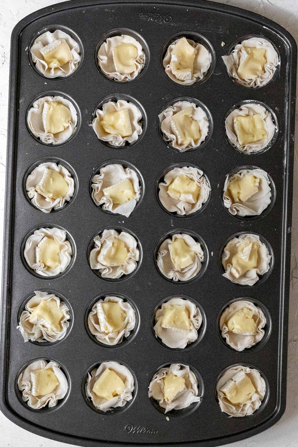 Raw phyllo cups in a mini muffin tin with a cube of brie in each one.