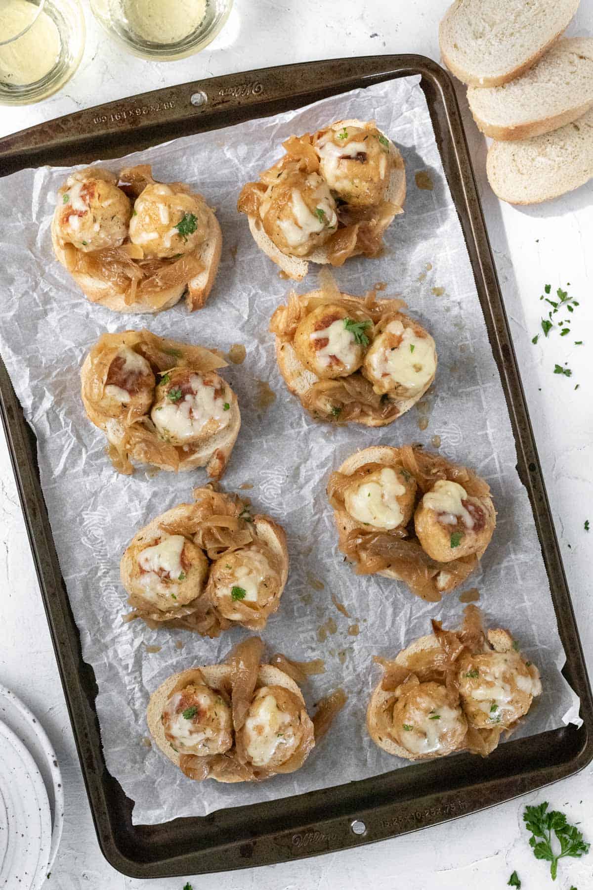 Toasted french bread topped with chicken meatballs, caramelized onions, and melted cheese lined up on a sheet pan..