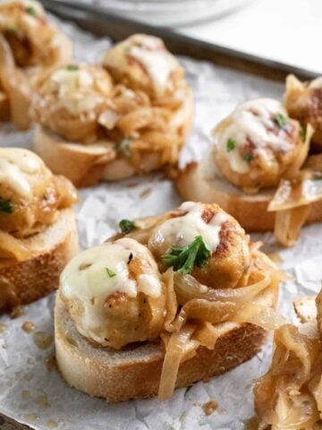 A closeup shot of 2 french onion chicken meatballs topped with melted cheese on a slice of toasted french bread.
