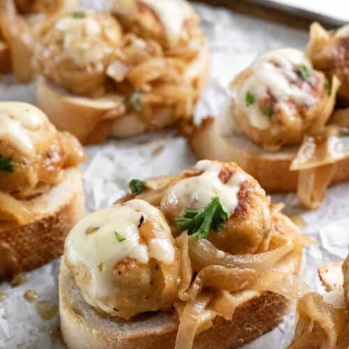 A closeup shot of 2 french onion chicken meatballs topped with melted cheese on a slice of toasted french bread.