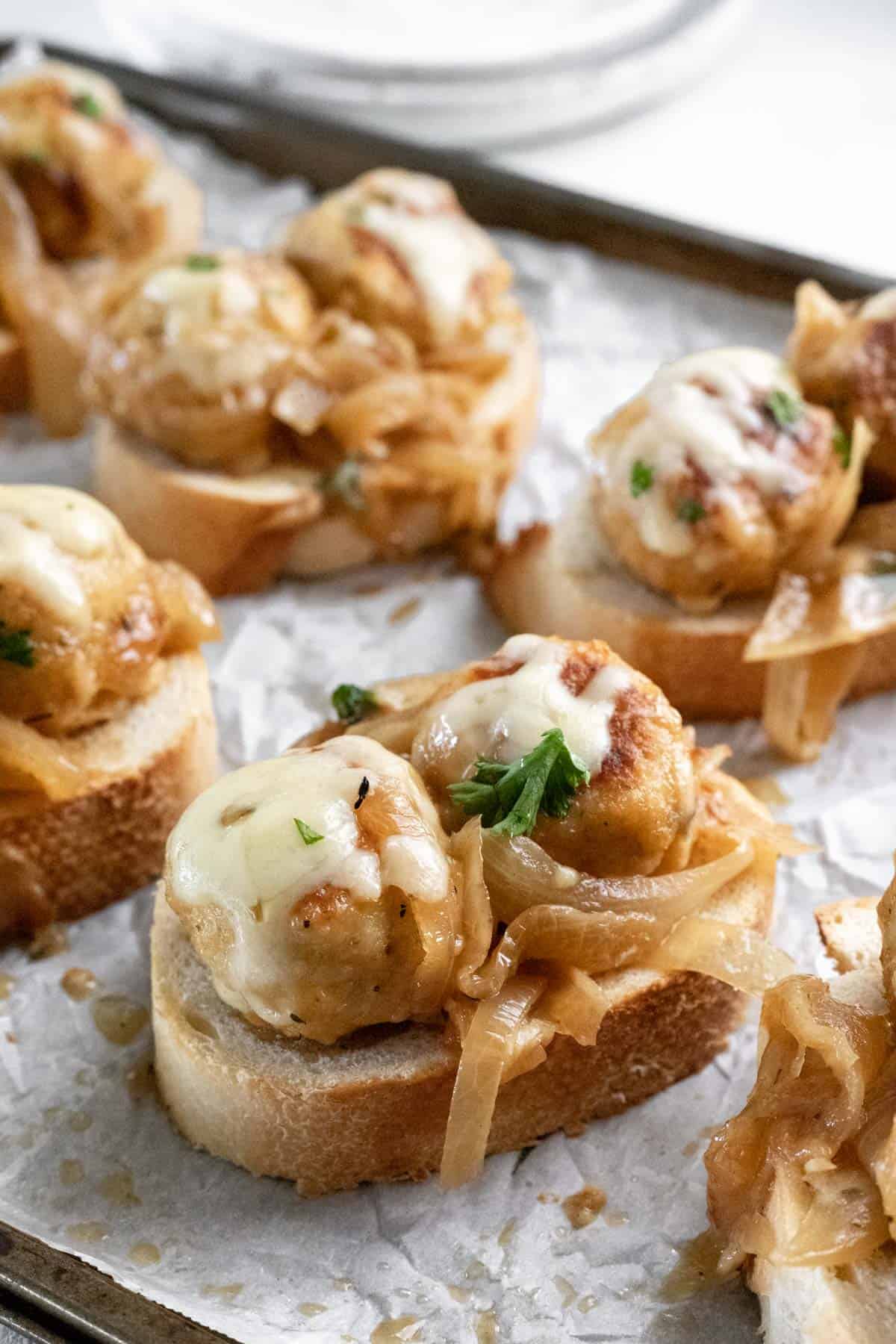 A closeup shot of 2 french onion chicken meatballs topped with melted cheese on a slice of toasted french bread.