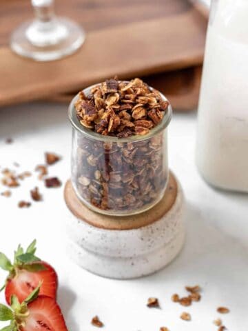 Homemade apple cinnamon granola in a glass jar with a jar of milk and fresh strawberries around it.