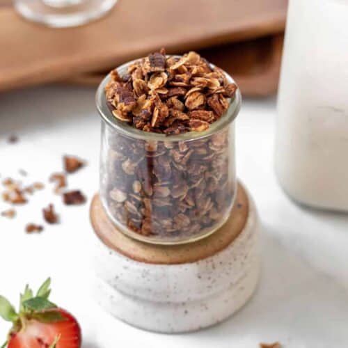 Homemade apple cinnamon granola in a glass jar with a jar of milk and fresh strawberries around it.