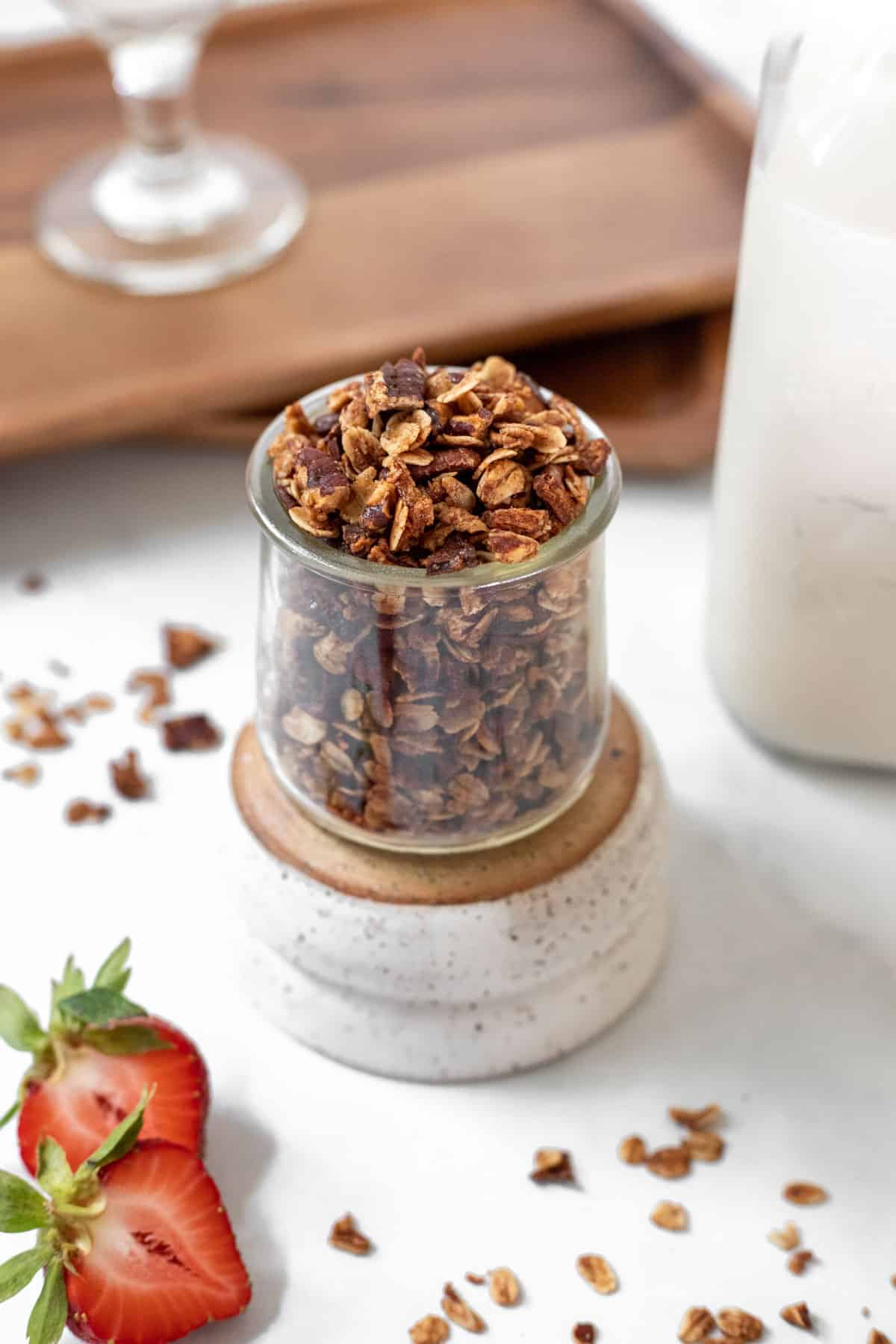 Homemade apple cinnamon granola in a glass jar with a jar of milk and fresh strawberries around it.