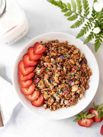 Birthday cake granola in a white bowl with sliced strawberries on top and a jar of milk next to it.