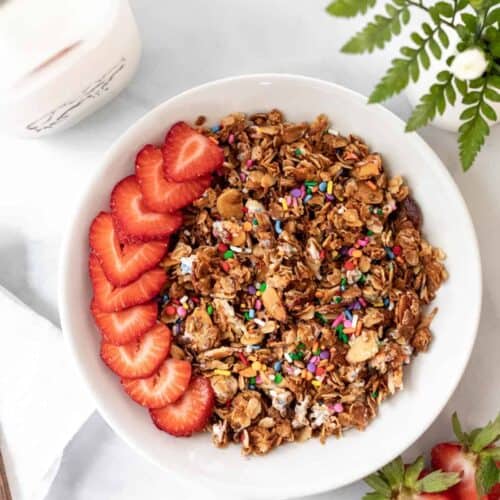 Birthday cake granola in a white bowl with sliced strawberries on top and a jar of milk next to it.