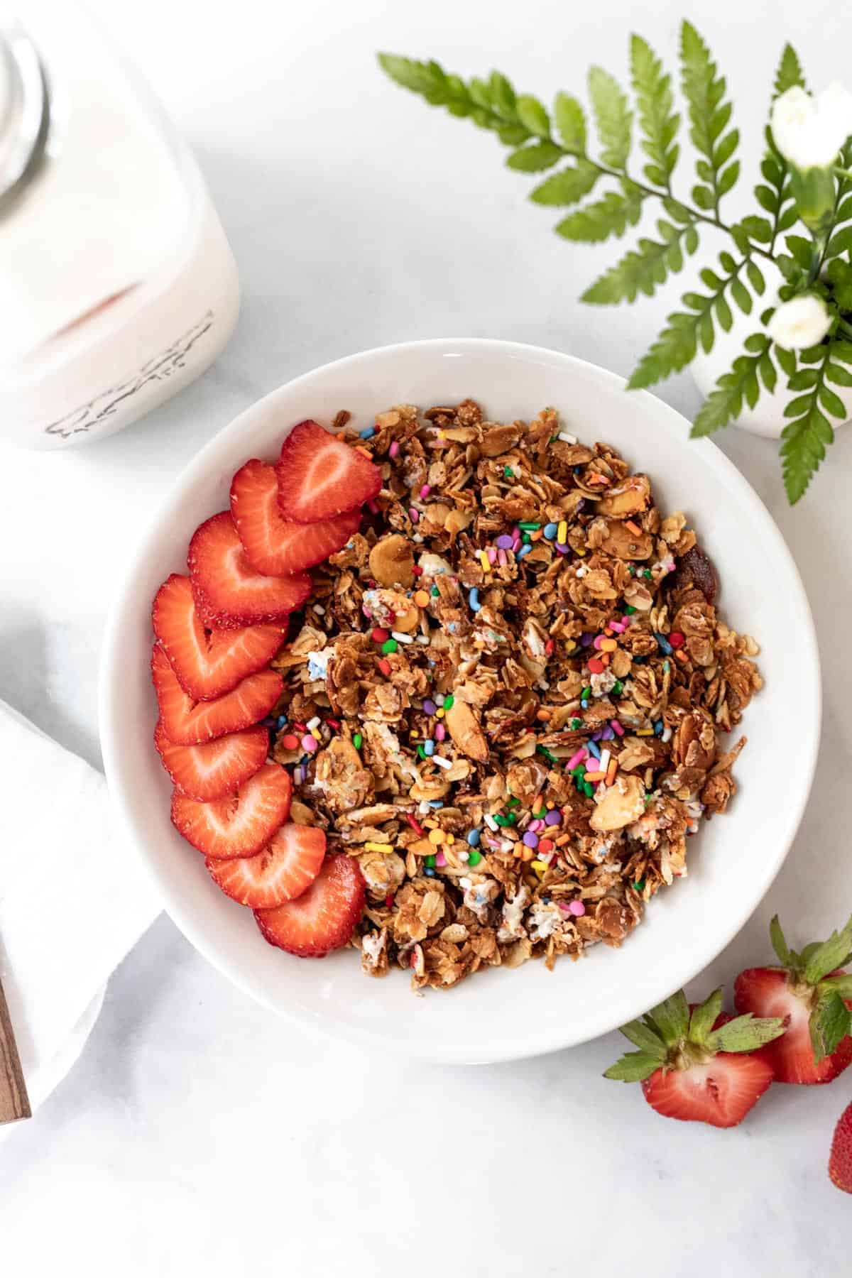 Birthday cake granola in a white bowl with sliced strawberries on top and a jar of milk next to it.