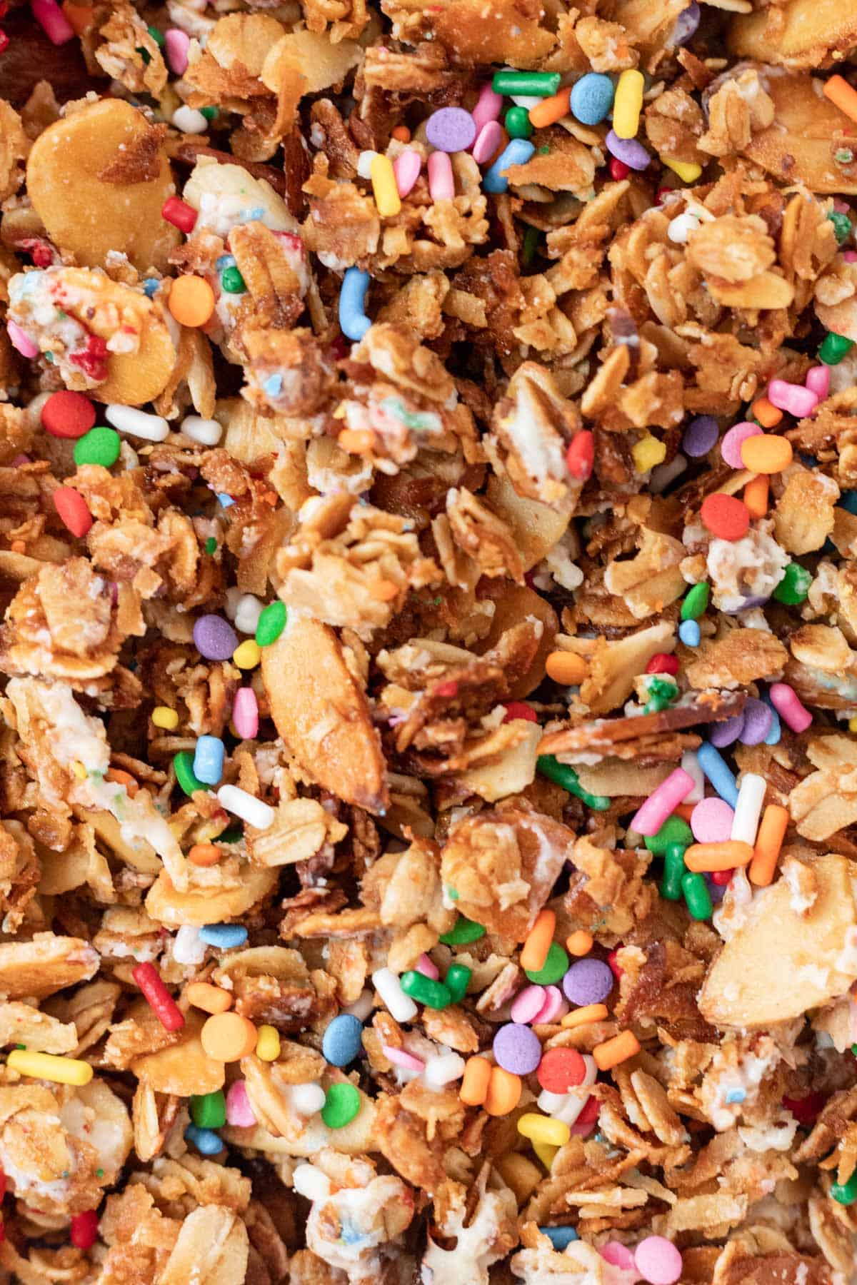 A closeup of baked granola with melted white chocolate and rainbow sprinkles.