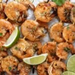 Grilled shrimp skewers garnished with limes and cilantro on a sheet pan.