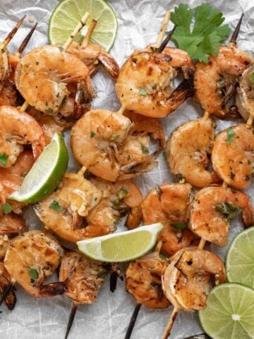 Grilled shrimp skewers garnished with limes and cilantro on a sheet pan.