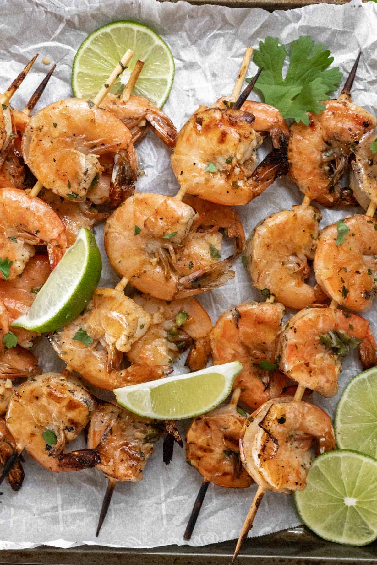 Grilled shrimp skewers garnished with limes and cilantro on a sheet pan.