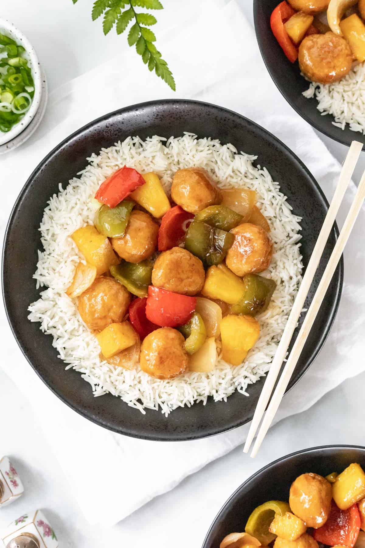 Sweet and sour pineapple chicken meatballs on a bed of white rice in a black bowl with chopsticks.