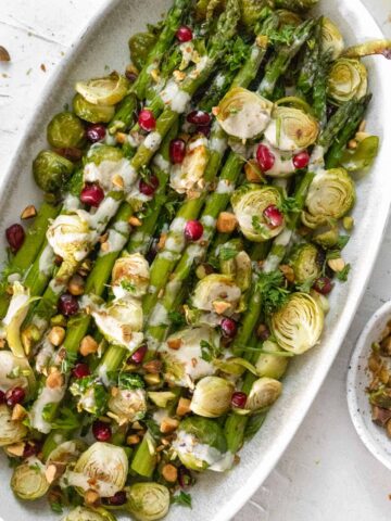 Roasted asparagus and brussels sprouts with a drizzle of lemon tahini sauce and a sprinkle of pistachios and pomegranate seeds on an oval platter.