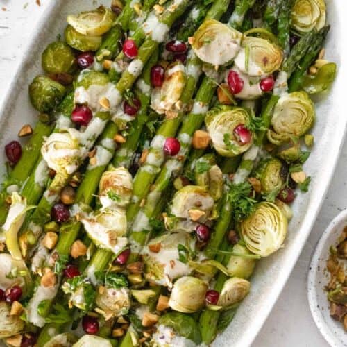 Roasted asparagus and brussels sprouts with a drizzle of lemon tahini sauce and a sprinkle of pistachios and pomegranate seeds on an oval platter.