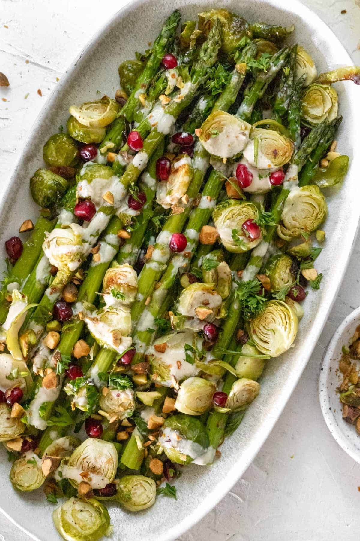 Roasted asparagus and brussels sprouts with a drizzle of lemon tahini sauce and a sprinkle of pistachios and pomegranate seeds on an oval platter.