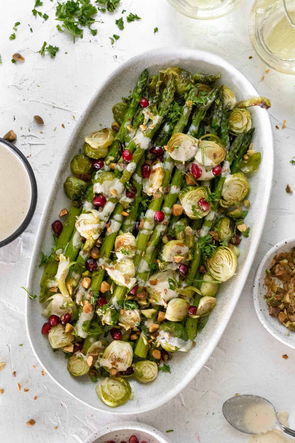 Roasted asparagus and brussels sprouts on an oval platter with bowls of extra sauce, pistachios, and pomegranate seeds around it.