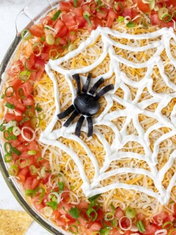 A closeup of a spider made out of black olives and a web made out of sour cream on top of Halloween taco dip.