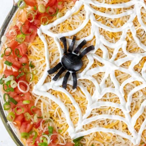 A closeup of a spider made out of black olives and a web made out of sour cream on top of Halloween taco dip.