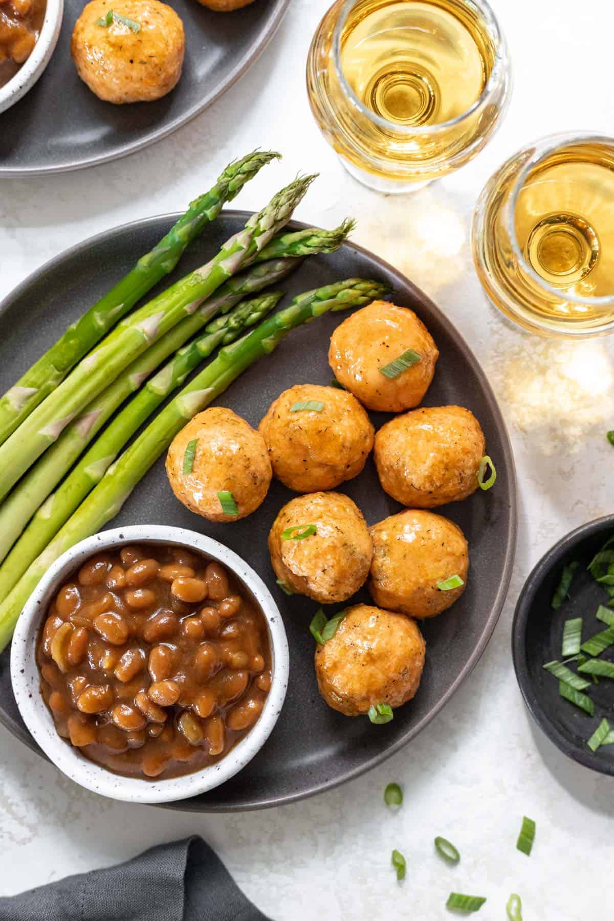 Honey bourbon glazed salmon meatballs on a gray plate with a side of asparagus and baked beans.