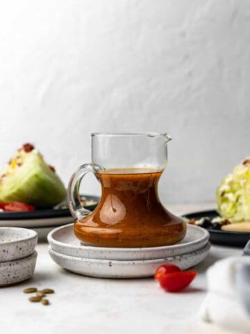 Pumpkin salad dressing in a small glass pitcher with wedges of salad and salad toppings next to it.