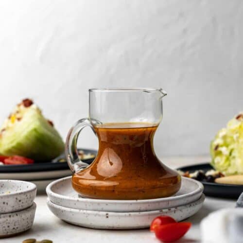 Pumpkin salad dressing in a small glass pitcher with wedges of salad and salad toppings next to it.