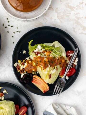 A wedge of iceberg lettuce on a blue plate with pumpkin vinaigrette, feta, tomatoes, chives, cranberries, and apples around it.