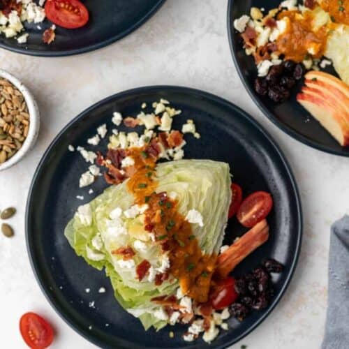 A wedge of iceberg lettuce on a blue plate with pumpkin vinaigrette, feta, tomatoes, chives, cranberries, and apples around it.