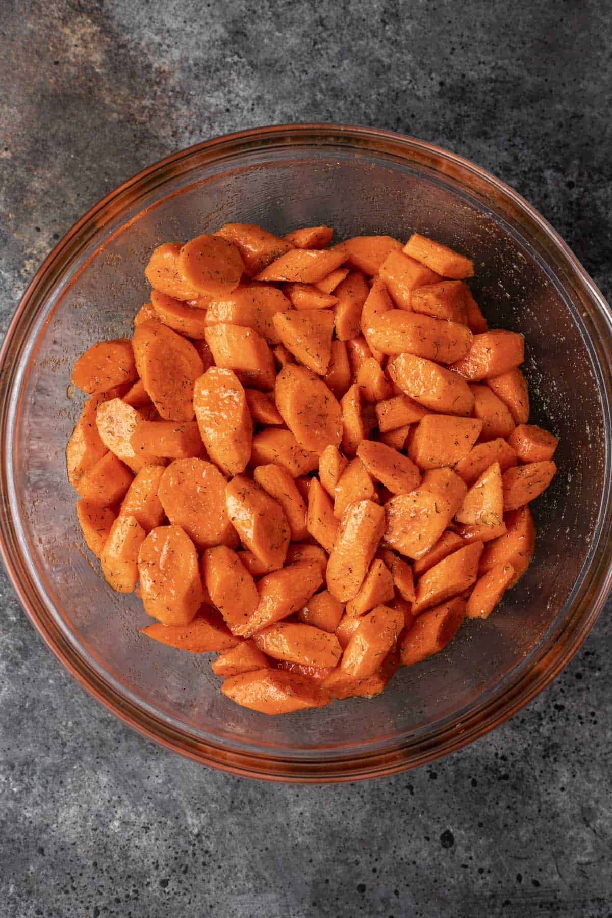 Carrots seasoned with olive oil, brown sugar, dill, and seasonings in a glass mixing bowl.