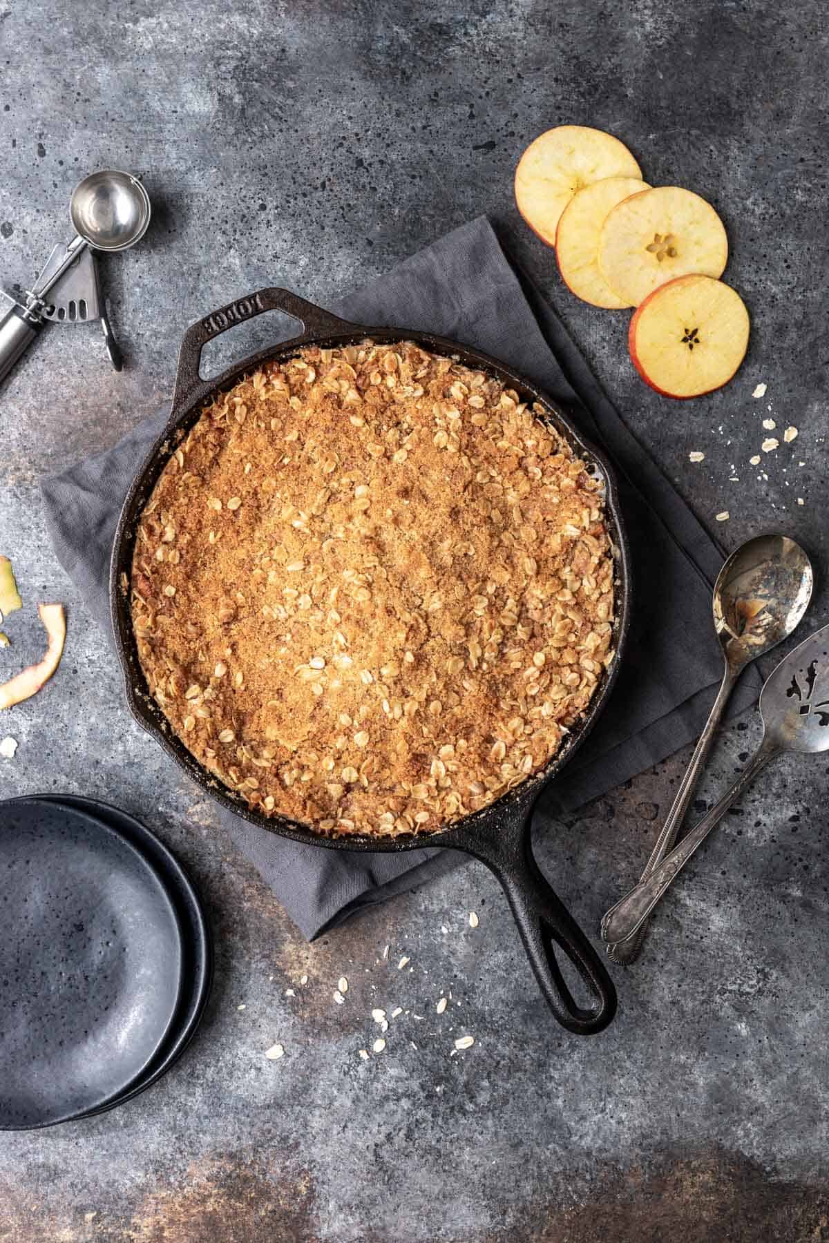 Golden brown apple crisp in a skillet with a side of sliced apples and serving plates.