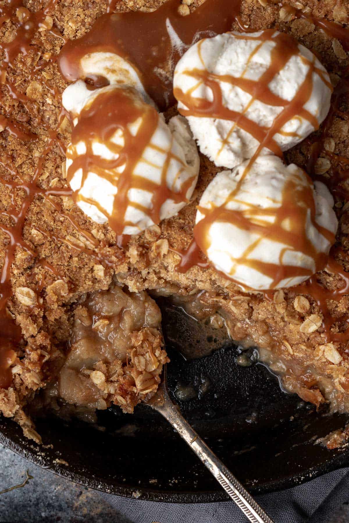 A large serving spoon scooping apple crisp topped with vanilla ice cream and caramel sauce from a skillet.